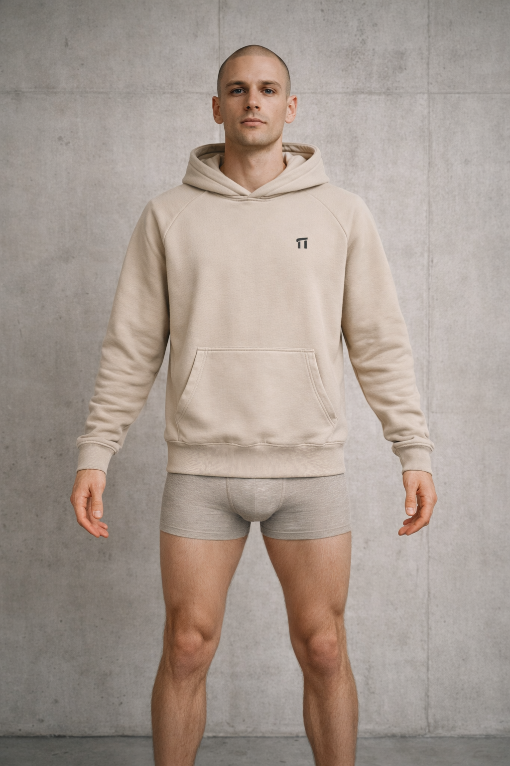 Man wearing a beige hoodie and matching underwear against a gray concrete wall.